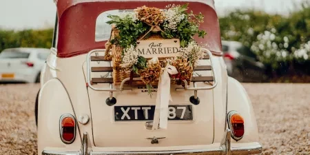 A photo of a vintage car with a Just Married sign attached to the back in a middle of a floral wreath.