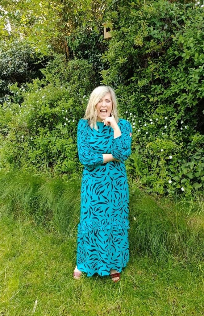 Sam is wearing a turquoises dress and is surrounded in beautiful greenery. Sam is just about to deliver a wedding ceremony.